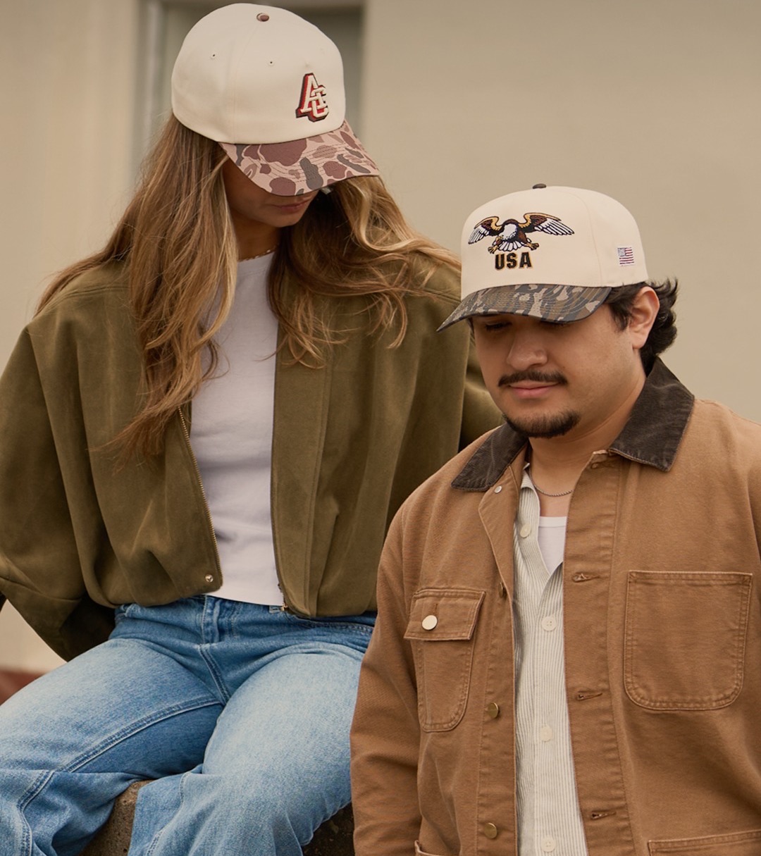 Two models in cream caps with camo brims sitting outdoors