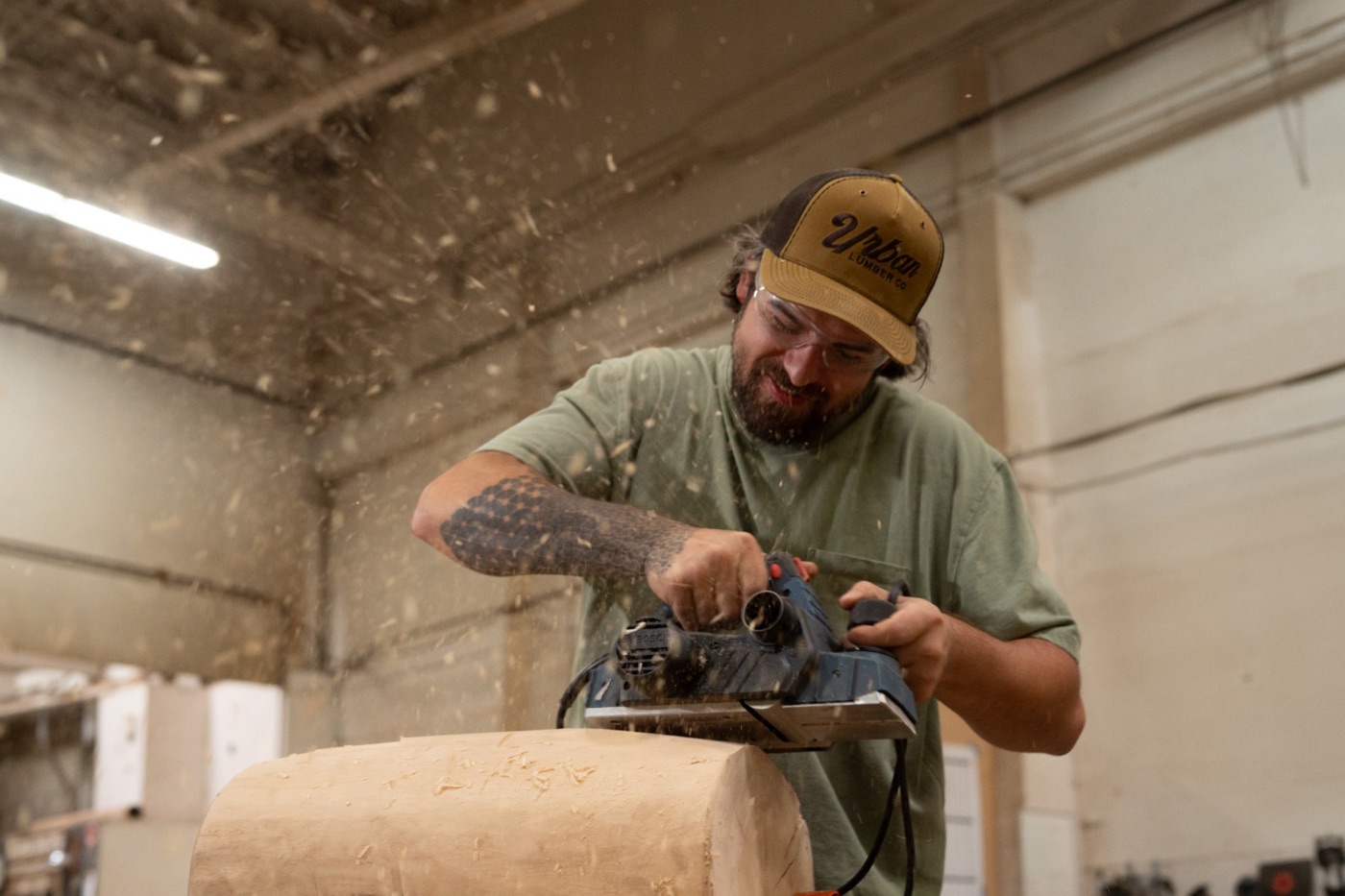 Woodworker in an Urban Lumber cap planing a log as shavings fly